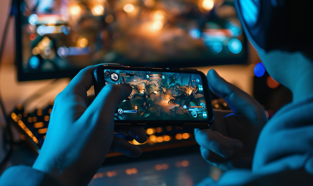 A person playing on a smartphone with a computer setup in the background